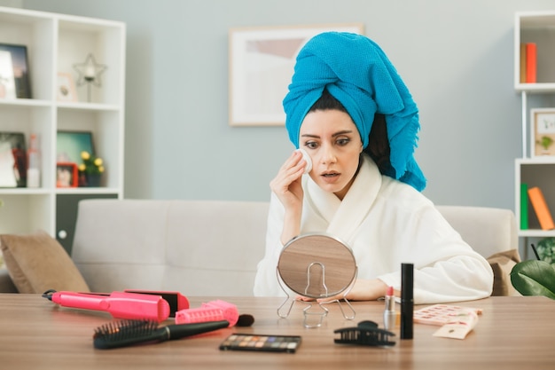 Young woman loooking at mirror applying tone-up cream with sponge wrapped hair in towel sitting at table with makeup tools in living room