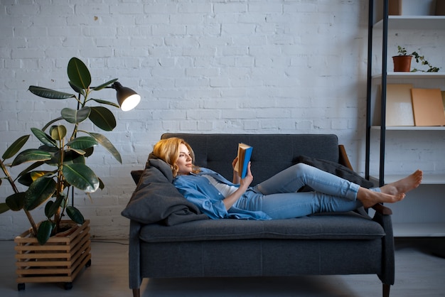 Young woman lying on cozy black couch and reading a book, living room in white tones