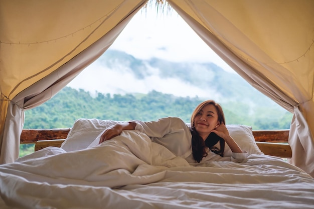 テントの外の美しい自然の景色と朝の白いベッドに横になっている若い女性