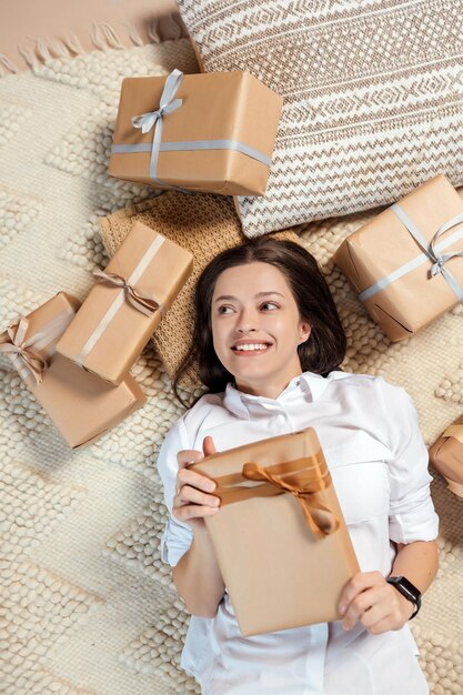 Young woman lying on floor with christmas presents and is holding in hand one handmade present packe...