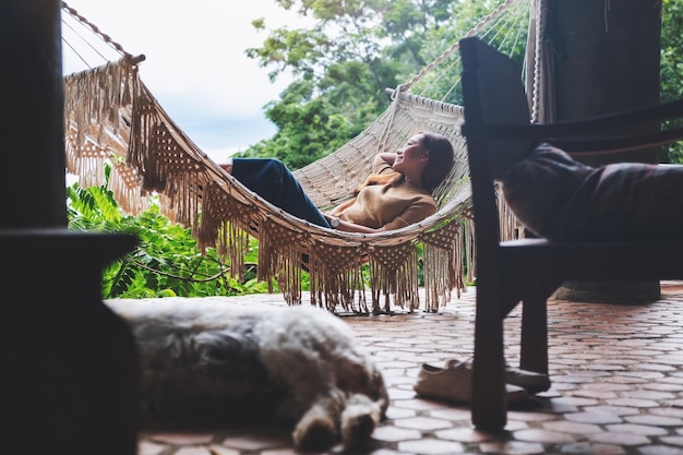 A young woman lying and relaxing on hammock with a dog sleeping on the floor