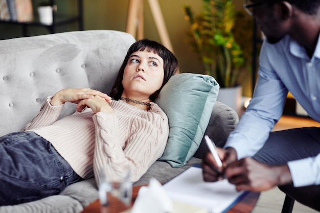 Young woman lying on sofa at therapy session confessing problems and looking up at her psychotherapist