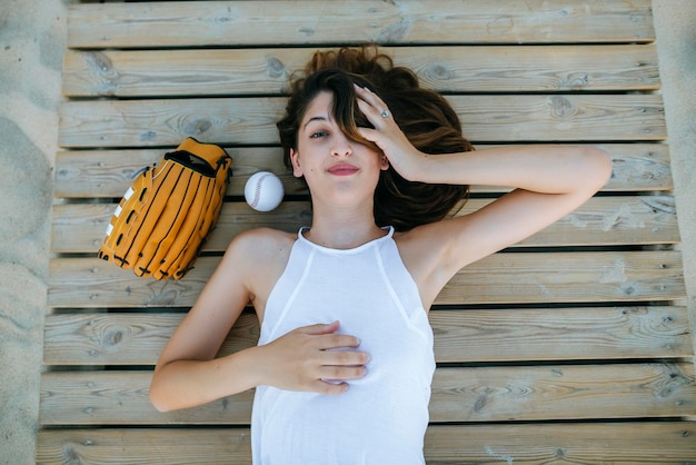 Молодая женщина лежит на деревянной дорожке рядом с мячом и бейсбольной перчаткой