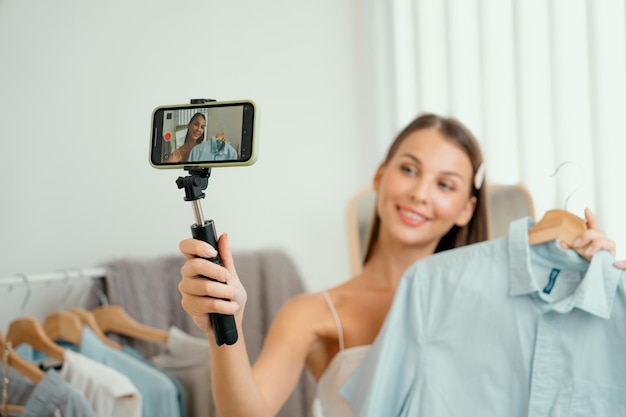 Young woman making fashion video content for social media Blogger smiles to camera using selfie stick and light ring while making persuasive online clothing sell to audience or followers Unveiling