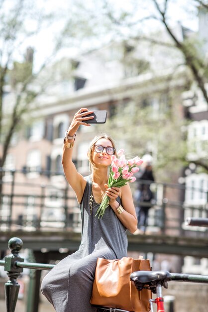 アムステルダム市の水路近くのフェンスに座っているピンクのチューリップの花束で自分撮り写真を作る若い女性