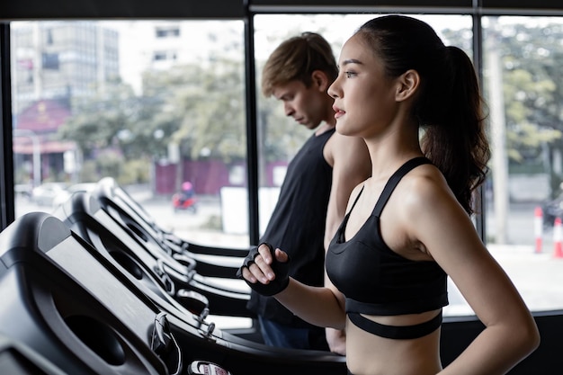 Young woman and man running side by side on modern electric treadmills at the gym. Pretty asian woman running on treadmill with fit young man on background. Young couple on electric treadmills.