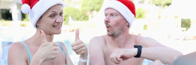 Young woman and man in santa claus hats in pool invites thumbs up gesture and smiles into laptop