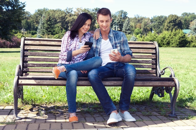 Молодая женщина и мужчина сидят на скамейке в парке и смотрят в смартфон