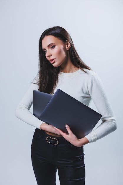 Young woman manager in a white shirt holds a portable laptop in her hands while standing on a gray background