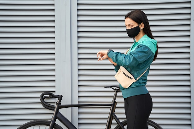 バイクの近くの通りのスマートウォッチで時間をチェックするマスクの若い女性。スポーツとライフスタイルのコンセプト