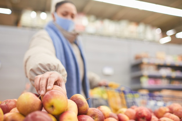 Young woman in mask choosing fresh apples while doing shopping in supermarket