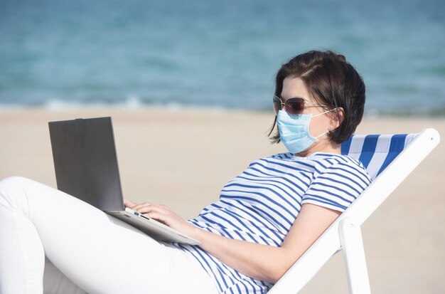 Giovane donna in maschera seduta con laptop su dispositivi di protezione della costa vuota durante il coronavirus covid19 concetto di distanza sociale