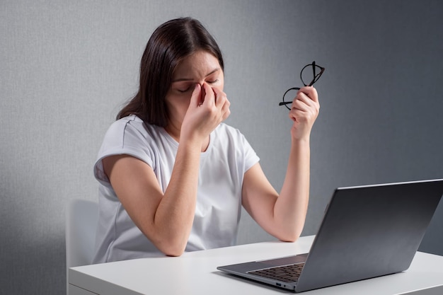 Young woman massaging the bridge of nose holding glasses in other hand sitting in front of a laptop