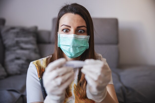 Young woman measuring temperature at home isolation, wearing protective mask and gloves.