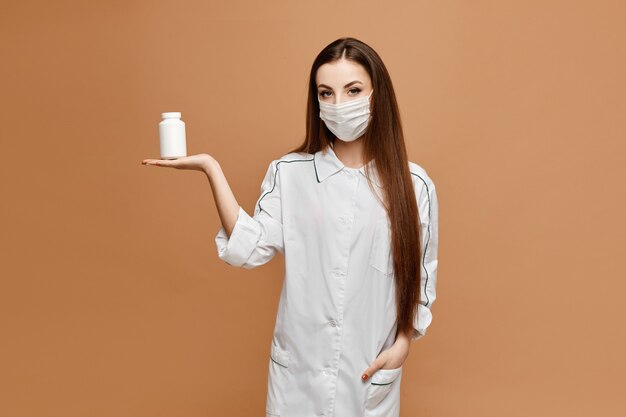 Young woman in medical clothes and protective mask posing with jar of pills. Female doctor keeps in hand pills. Virus and flu protection pills concept.