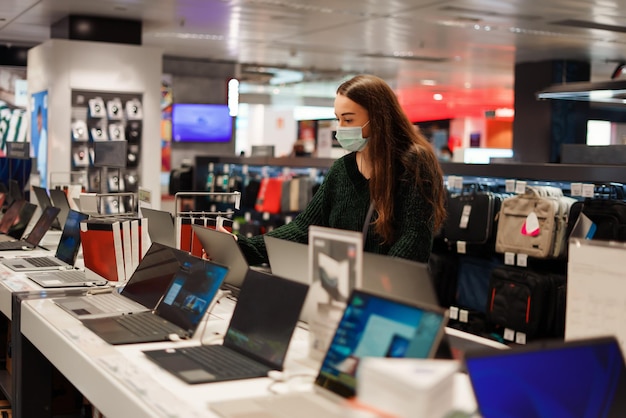 Young woman in medical face mask choosing laptop in tech store during covid pandemic