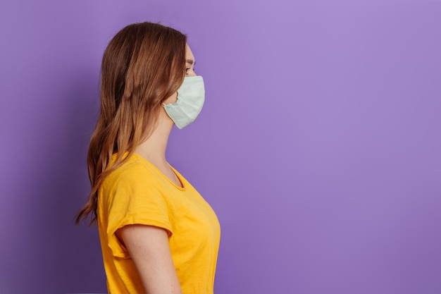 Young woman in medical face mask stands in profile isolated on lilac background in studio