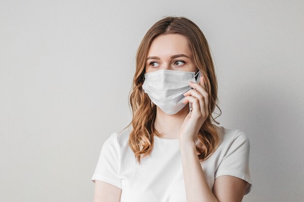 Young woman in a medical mask calls on the phone and stands over the wall, looks away