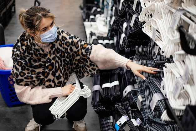 Young woman in medical mask crouches down and selects set of plastic hangers for organizing dressing room space in shopping center. New normal concept. Shopping in epidemic. Order in house. Lifestyle.