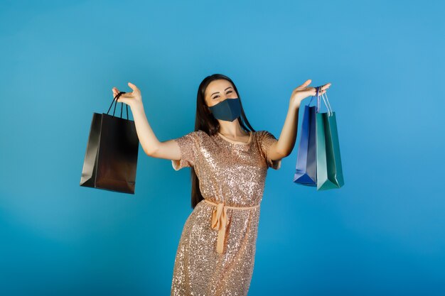 Young woman in medical mask holding gift bags on blue background