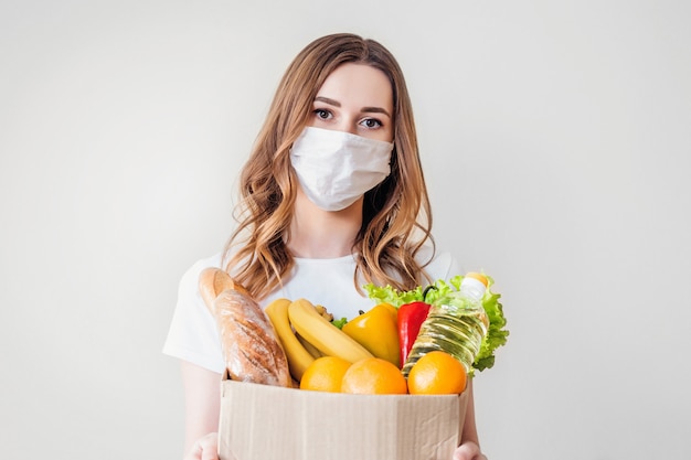 Young woman in medical mask holds a paper cardboard box with food, fruits and vegetables, baguette, lettuce over grey background, home delivery, coronovirus, quarantine, stay home concept, copy space