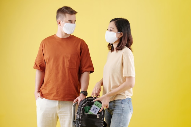 Young woman in medical mask putting smartphone with covid vaccine passport in backpack when talking ...