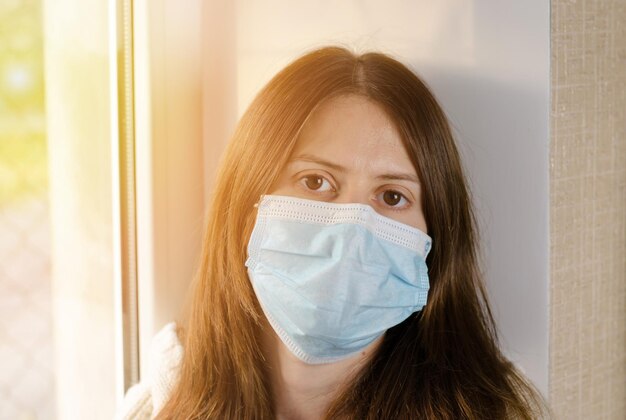 A young woman in a medical mask sits by the window in the sun self-isolation concept