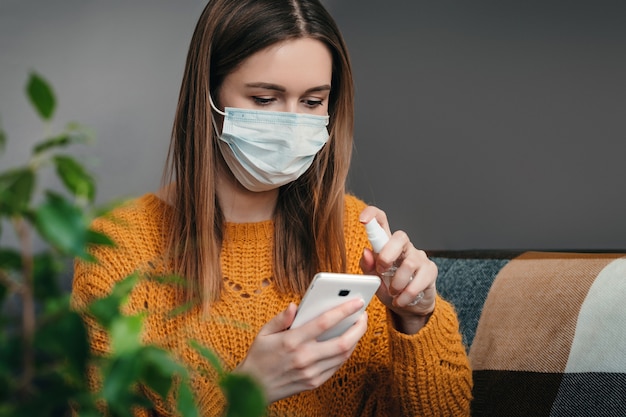 Una giovane donna in una maschera medica si siede a casa in isolamento di quarantena e lava il suo telefono cellulare con una soluzione di spray antisettico batteri coronovirus parete scura da vicino