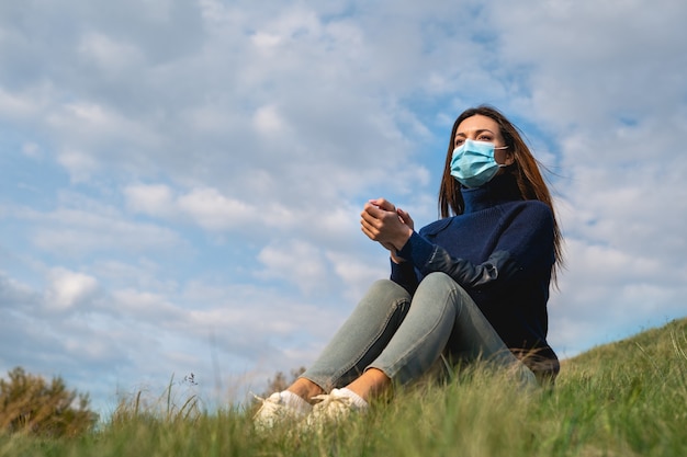 The young woman in medical mask sitting on the green hill