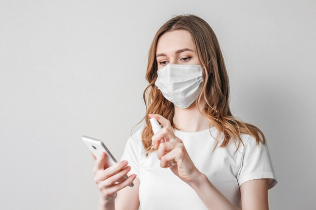 A young woman in a medical mask washes her mobile phone with a solution of antiseptic, spray