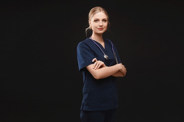 Una giovane donna in uniforme medica e con uno stetoscopio in posa con le mani giunte sul buio