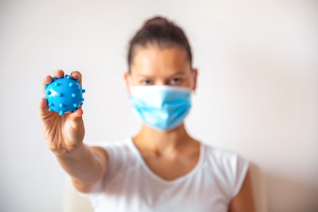Молодая женщина в медицинской маске с синим пластиковым шаром в виде вируса в руке на белом фоне, медицинская концепция, концепция остановки коронавируса COVID-19