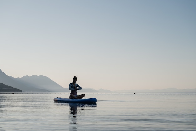 Young woman meditating in lotus position with her hand joined in front of her chest, sitting on sup board floating on calm morning water.