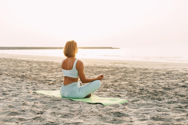 young woman meditating in pose of lotus on beach near the sea at sunset in summer rear view