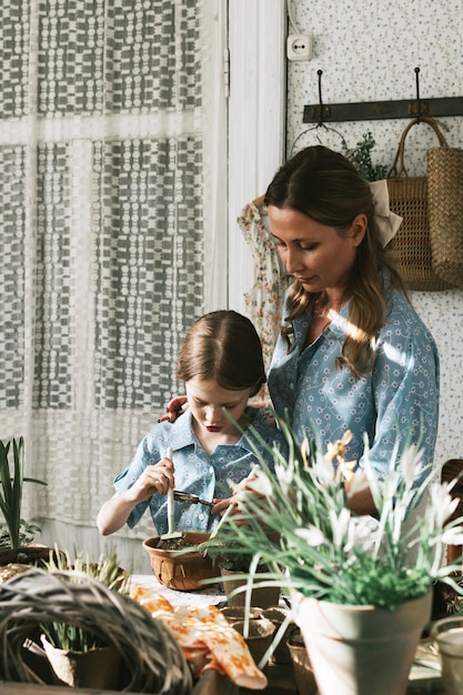 写真 若い女性と彼女の小さな娘は家族の見た目でドレスを着て春のテラスで花を植えています家庭の庭園で苗が育つ田舎の家ベランダ母性