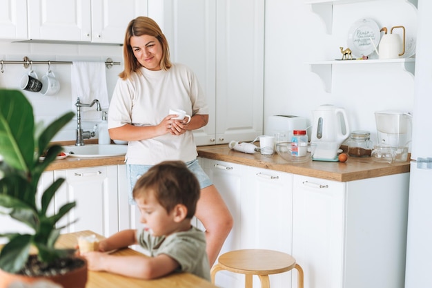 自宅のキッチンのテーブルで朝食をとっている彼女の幼児の男の子の息子のように見える若い女性の母親