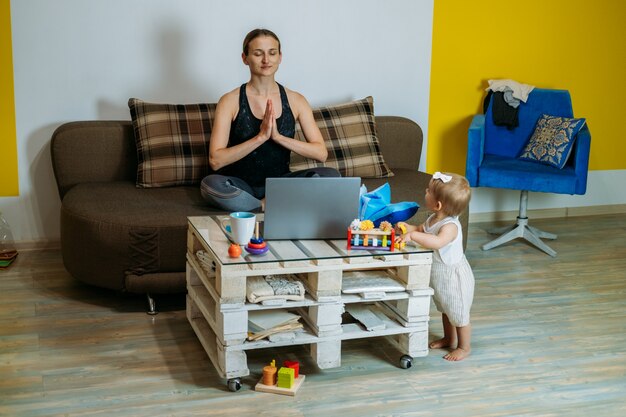 Young woman mother sit on sofa with her baby toddler daughter at home doing yoga exercises watching