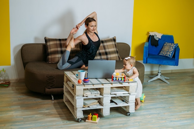 Young woman mother sit on sofa with her baby toddler daughter at home doing yoga exercises watching