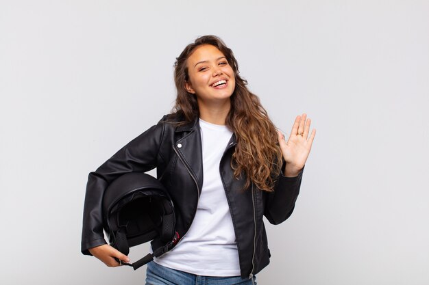 Young woman motorbike rider smiling happily and cheerfully, waving hand, welcoming and greeting you, or saying goodbye