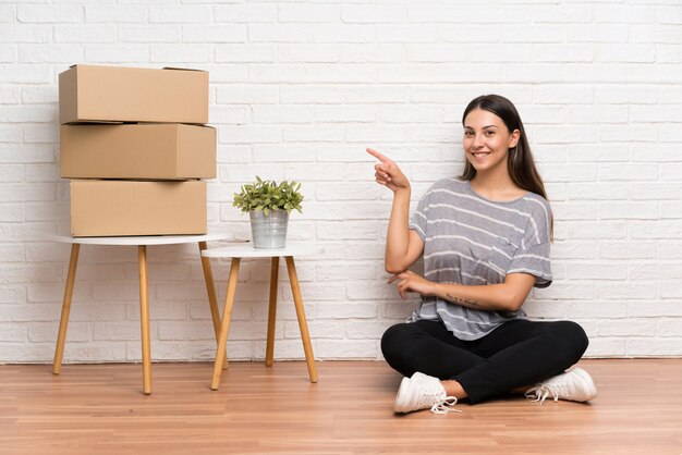 Young woman moving in new home among boxes pointing finger to the side