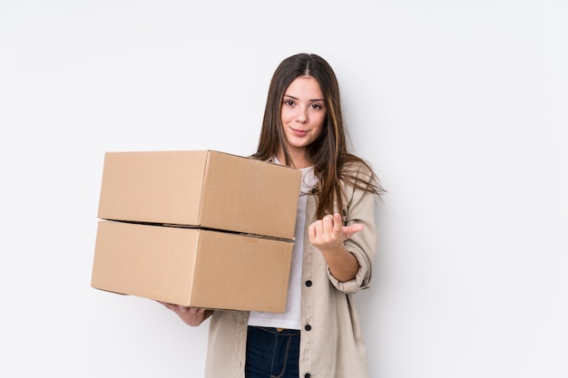 Young woman moving to a new home pointing with finger at you as if inviting come closer.