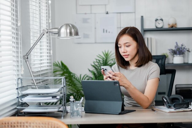 A young woman multitasking in a home office using a smartphone and tablet surrounded by modern office supplies and decor