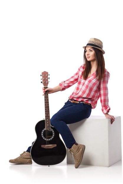 Музыкант молодой женщины с гитарой сидя на кубе и откинувшись назад. Белый фон.