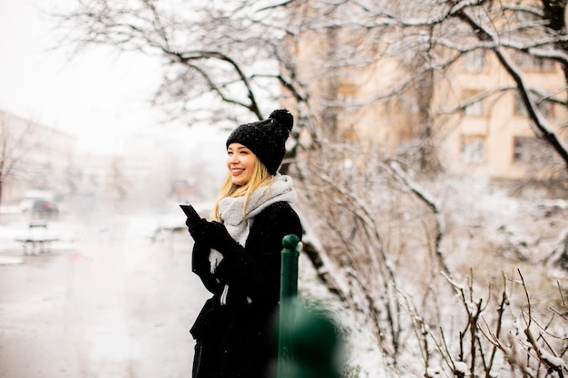 Молодая женщина в теплой одежде наслаждается снегом и пользуется мобильным телефоном