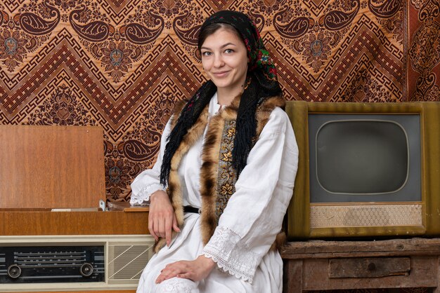 young woman in national clothes standing in room with vintage wallpaper and interior with tv and radio