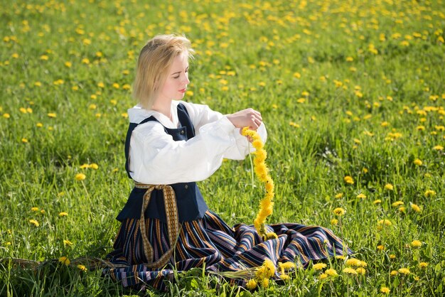 Young woman in national clothes wearing yellow dandelion wreath in spring field springtime