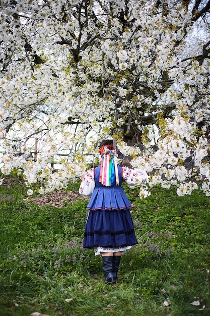 Young woman in national ukrainian costume smiling young lady in\
traditional clothes stay near blooming tree peace in ukraine