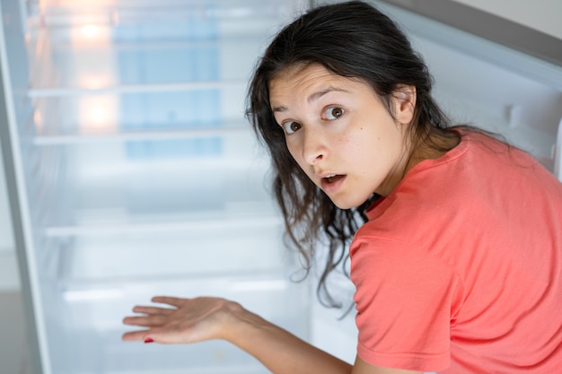 食べ物のない空の冷蔵庫の近くの若い女性