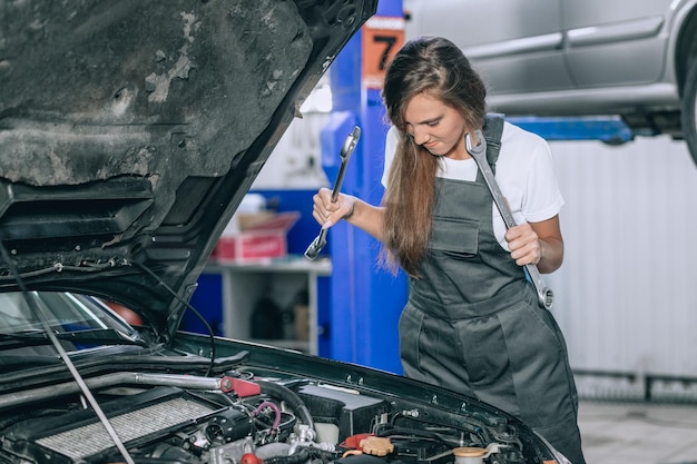 Young woman near the open hood of a black car in the garage. The girl is dressed in black jumpsuit and white t-shirt, she holds up the wrenches and lowers her gaze.