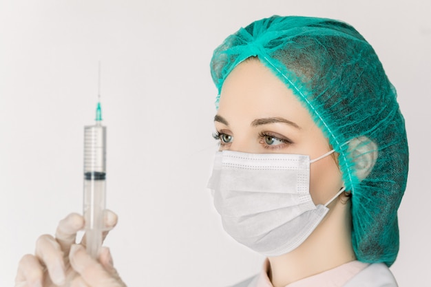 Young woman nurse or doctor in white lab coat, cap and mask, with syringe in hands, prepares syringe for vaccination, isolated on white background. Medical and health care concept